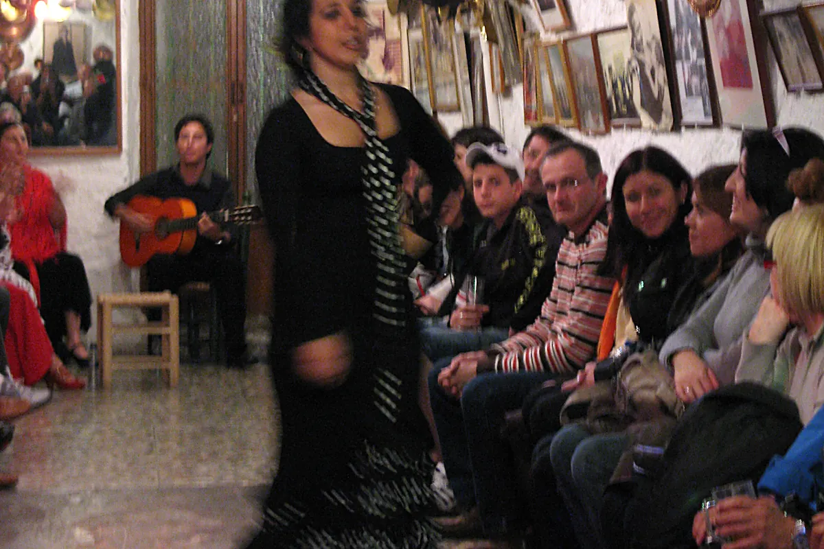 A dancer in black with a polka-dot scarf passes a seated guitarist as spectators watch from the cave's side benches
