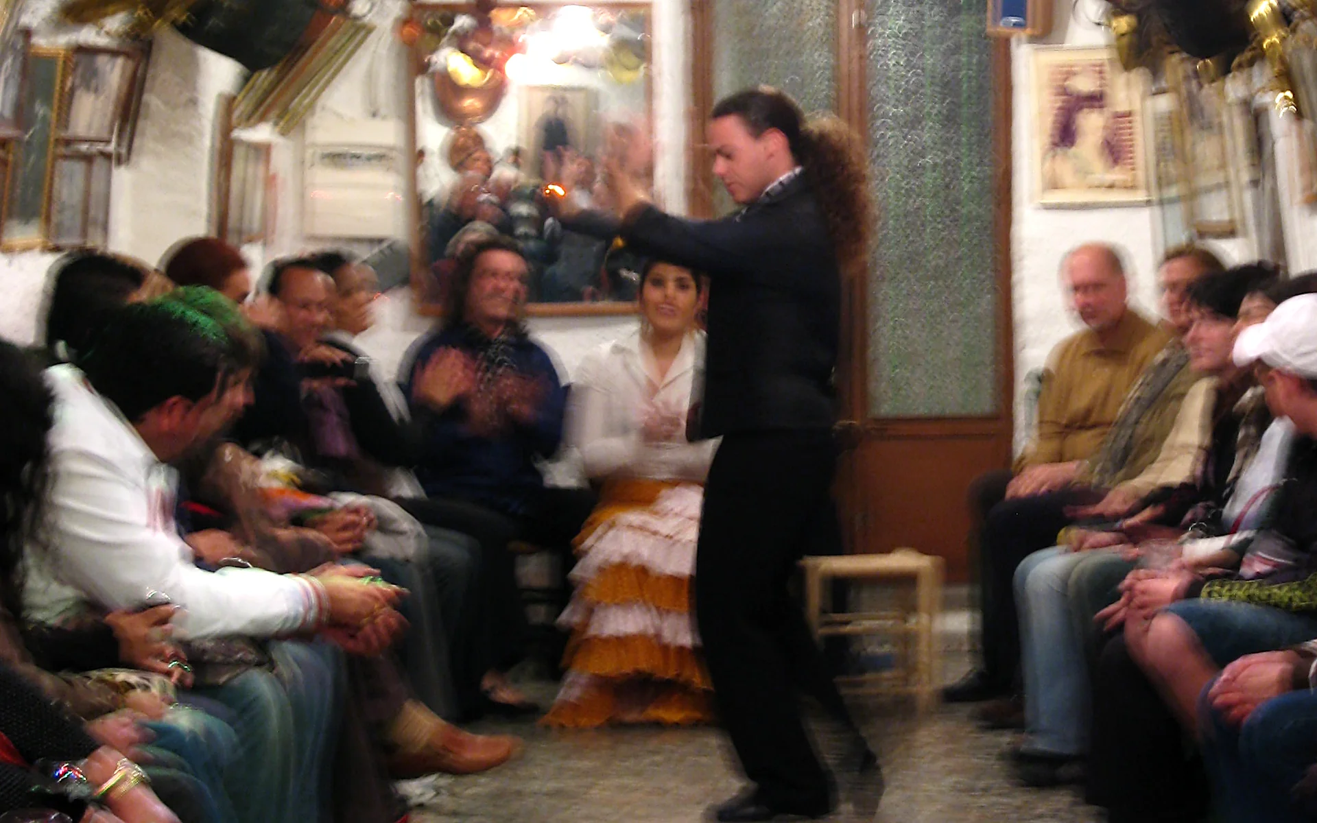A dancer in black with a ponytail performs in the aisle of a whitewashed cave, a seated woman in a ruffled skirt clapping behind him and spectators along both walls; a photograph from April 2009 that Wikimedia Commons labels Cueva de la Rocío, so today's cast and details will differ